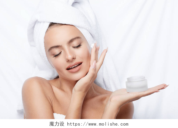在白色的床单前一个美丽的女人在护肤漂亮的微笑的年轻女子使用奶油在白色背景, 头上的毛巾, 美容照片, 护肤, 水疗理念